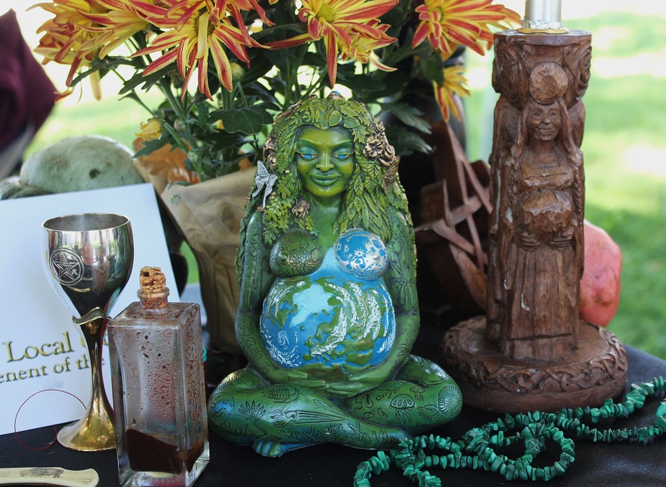 A Wiccan-style gathering of artifacts including a statue of a seated green goddess, her pregnant belly painted as the earth; mums; a chalice; a string of green beads; a stoppered rectangular prism bottle; and a candleholder appearing to be carved of wood, again of a pregnant woman with hands holding her belly.