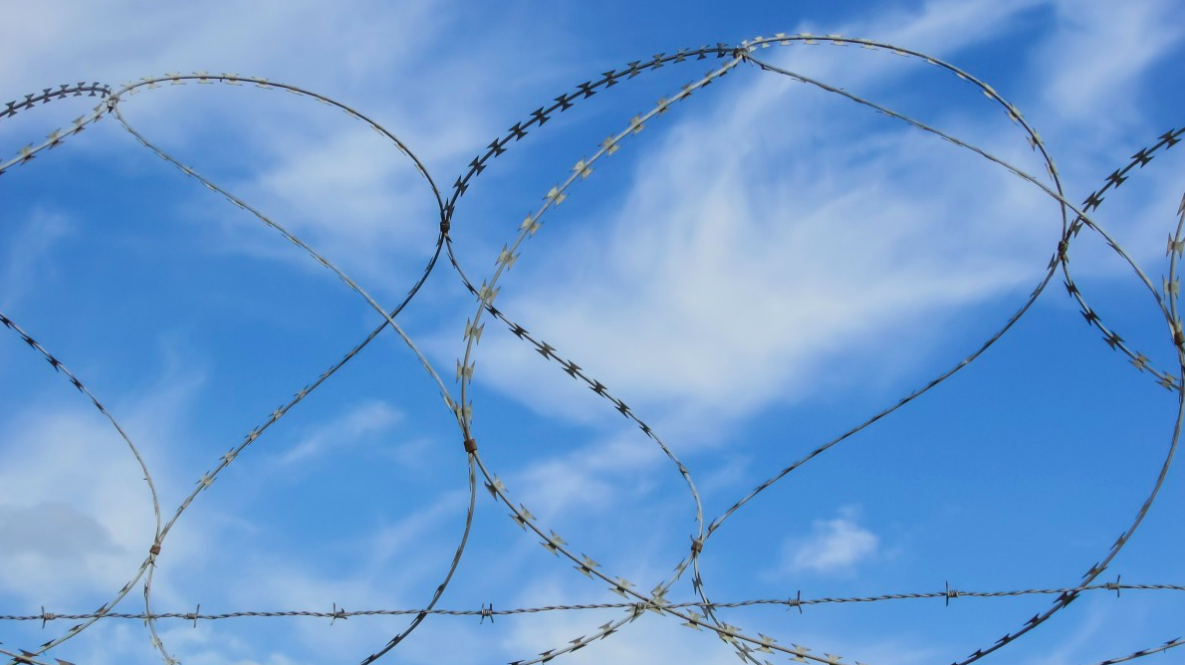 Blue sky and white cirrus clouds as viewed through coiled razor wire atop a barbed-wire fence.